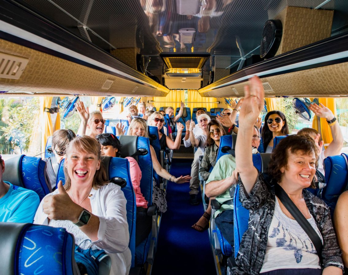 A front-view shot of a large multi-ethnic group of tourists celebrating on a coach bus, they are smiling and raising their arms with excitement, they are ready to begin their journey.